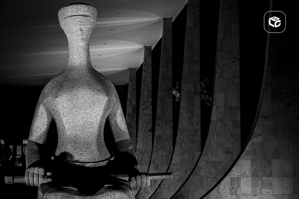 Fotografia em preto e branco de uma grande escultura estilizada representando uma figura sentada segurando um objeto horizontal nas mãos. A escultura ocupa o primeiro plano da imagem e apresenta textura metálica brilhante. Ao fundo, há uma sequência de colunas altas e curvas de um edifício monumental, iluminadas por luzes que criam padrões geométricos no teto. O ambiente é noturno, com forte contraste entre áreas iluminadas e sombras profundas. No canto superior direito há um pequeno logotipo branco dentro de um quadrado de cantos arredondados.