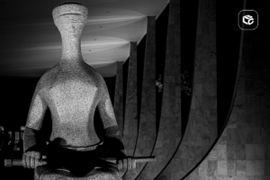 Fotografia em preto e branco de uma grande escultura estilizada representando uma figura sentada segurando um objeto horizontal nas mãos. A escultura ocupa o primeiro plano da imagem e apresenta textura metálica brilhante. Ao fundo, há uma sequência de colunas altas e curvas de um edifício monumental, iluminadas por luzes que criam padrões geométricos no teto. O ambiente é noturno, com forte contraste entre áreas iluminadas e sombras profundas. No canto superior direito há um pequeno logotipo branco dentro de um quadrado de cantos arredondados.